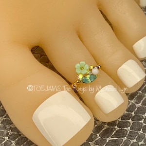 May include: A gold-toned toe ring featuring a cluster of decorative elements. The ring is adorned with a light green flower, a pale pink bead, and an oval-shaped teal gem. The text "TOEJAMS Toe Rings by Michelle Lyn" is visible.