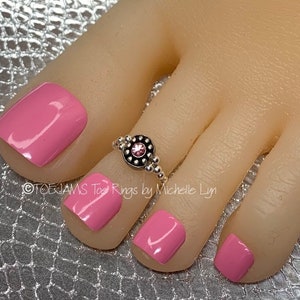 May include: A silver toe ring with a pink rhinestone in the center. The ring is on a foot with pink nail polish.
