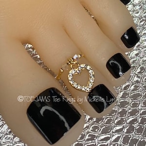 May include: A gold-colored toe ring with a heart-shaped pendant encrusted with clear crystals. The ring is worn on a foot with black nail polish. The text "TOEJAMS Toe Rings by Michelle Lyn" is visible.
