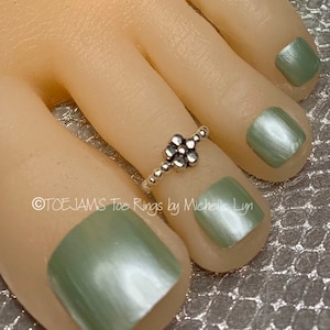 May include: A silver toe ring with a flower design is worn on a foot with green nail polish. The ring is made of small, round beads and a flower-shaped centerpiece. The text "TOEJAMS Toe Rings by Michelle Lyn" is visible.