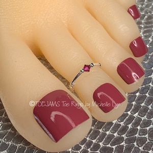 May include: A silver toe ring with a small pink gemstone. The ring is on a model's toe with mauve nail polish.
