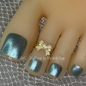 May include: A gold-toned toe ring featuring a central bow design encrusted with small clear stones. The ring is positioned on a toe with metallic blue nail polish. The text "TOEJAMS Toe Rings by Michelle Lyn" is visible on the image.