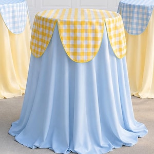 May include: Three round cocktail tables covered with floor-length fabric. The center table features a light blue base cloth with a yellow and white gingham scalloped topper. The two side tables have pale yellow bases with light blue gingham toppers.