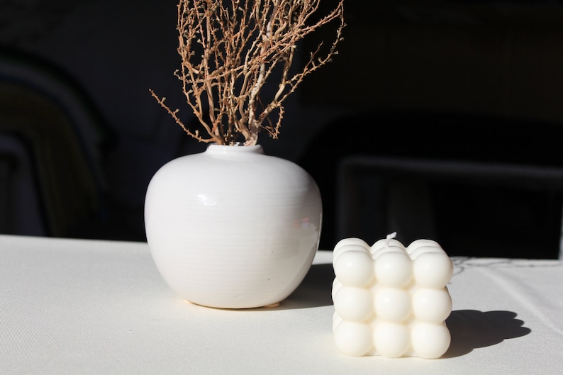 May include: A white ceramic vase with a dried branch inside, sitting on a white surface next to a white candle with a textured, bubble-like design.
