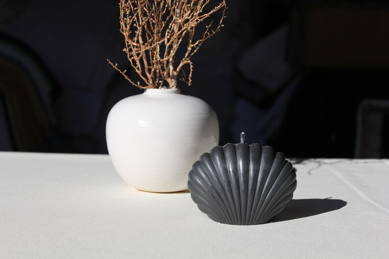 K&ouml;nnte beinhalten: Eine wei&szlig;e Keramikvase mit getrockneten Zweigen und einer schwarzen muschelf&ouml;rmigen Kerze auf einer wei&szlig;en Oberfl&auml;che.
