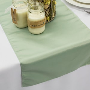 May include: Sage green satin table runner with three clear glass jars with white candles. The jars have brown paper tags with the text "Bride" and "Mr. & Mrs."