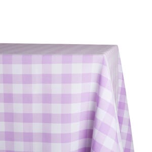 May include: A corner view of a lavender and white checkered tablecloth. The fabric is draped over a table, with the pattern clearly visible. The gingham pattern is made up of evenly sized squares.