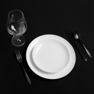 Puede incluir: Un plato blanco para cenar con un plato blanco m&aacute;s peque&ntilde;o dentro, un tenedor negro, una cuchara negra y un cuchillo negro sobre una superficie negra. Un vaso de vino de vidrio transparente est&aacute; a la izquierda del plato.