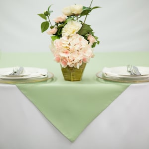 May include: A green tablecloth with a white tablecloth underneath. Two place settings with white plates, silver forks and knives, and clear glass glasses are on the table. A vase with pink and white flowers is in the center of the table.