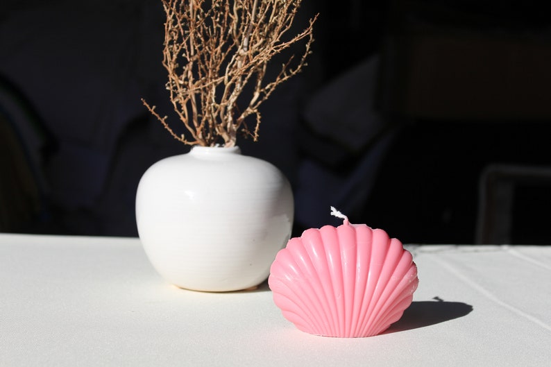 K&ouml;nnte beinhalten: Eine rosa Muschel-f&ouml;rmige Kerze auf einer wei&szlig;en Oberfl&auml;che neben einer wei&szlig;en Vase mit getrockneten Zweigen.