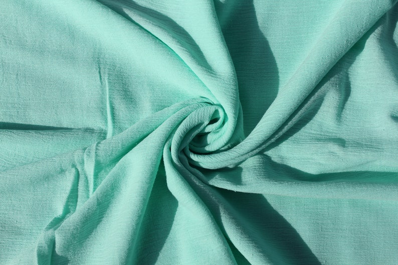 Puede incluir: Una tela suave de color verde claro con una textura arrugada. La tela est&aacute; doblada y drapeada en forma de espiral.