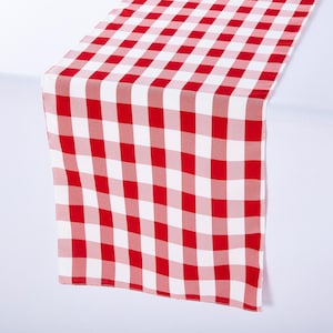 May include: A red and white checkered table runner. The runner is made of a fabric with a classic gingham pattern. The red and white squares create a bold, eye-catching design. This table runner is perfect for adding a touch of rustic charm to any dining table.