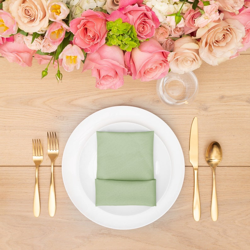 May include: A white dinner plate with a green folded napkin on top of a smaller white plate. The plate is set on a wooden table with gold silverware and a glass of water.