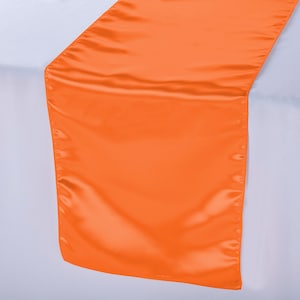 May include: A bright orange satin table runner. The runner is laying flat on a white tablecloth.