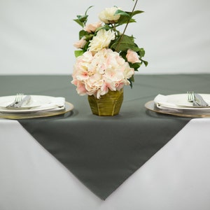 May include: A gray tablecloth with two place settings, each with a white plate, a gold charger plate, and a fork and knife. A vase with pink and white flowers sits in the center of the table.