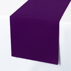 May include: A deep purple table runner draped over a white surface. The runner is made of a solid, smooth fabric and is displayed at an angle, showcasing its color and texture. The contrast between the purple and white creates a visually appealing presentation.