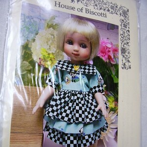 以下が含まれることがあります： 青いチェック柄のドレスと白いベレー帽をかぶった、25cmのドール。金髪で青い目。ドールは木製の台の上に立っており、背景には花が咲いています。画像の下部には「10" Ann Estelle Ruffled Dress and Beret」というテキストが印刷されています。