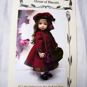 Puede incluir: Una muñeca de 33 cm vestida con un abrigo, un sombrero y un manguito burdeos. La muñeca tiene el pelo castaño y los ojos azules. El atuendo está ribeteado con piel marrón y tiene borlas burdeos.