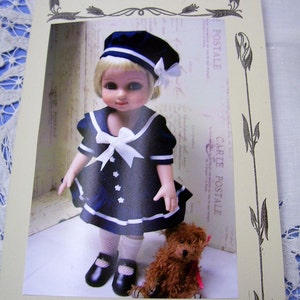 May include: A 10-inch doll wearing a blue and white sailor dress with a white bow and a blue and white hat. The doll is holding a brown teddy bear. The image is from House of Biscotti.