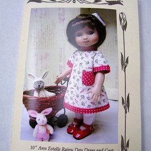 May include: A 10-inch Ann Estelle doll wearing a white and red dress with a red polka dot pattern and a red bow in her hair. The dress has a pattern of umbrellas. The doll is holding a white bunny in a basket.