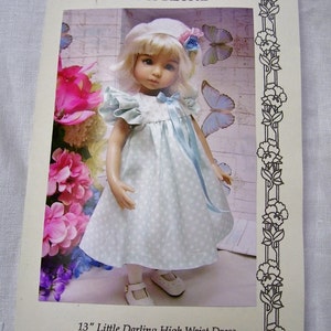 Puede incluir: Una muñeca de 33 cm vestida con un vestido azul claro con lunares blancos, un sombrero blanco con un lazo azul y zapatos blancos. La muñeca está de pie frente a un fondo floral.