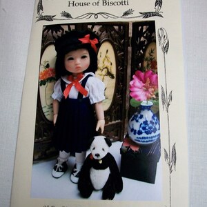 May include: A pattern for an 8-inch Ten Ping Asian doll wardrobe. The pattern features a doll wearing a blue dress with a red bow, white blouse, and black shoes. The doll is standing next to a black and white panda bear.