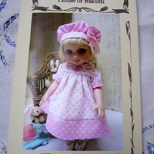 Può includere: Una bambola di 25 cm che indossa un vestito a pois rosa e bianco, un cappello bianco e scarpe bianche. La bambola tiene un peluche. L'immagine è di House of Biscotti.