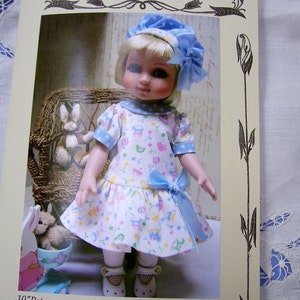 Peut inclure: Une poupée de 25 cm portant une robe à taille basse bleu et blanc avec un nœud bleu, un chapeau bleu à pois blancs et des collants blancs. La poupée est assise dans un fauteuil en osier avec un lapin en peluche à côté d'elle. L'image provient d'un livret de patrons pour une poupée nommée "Patsy" par Ann Estelle.