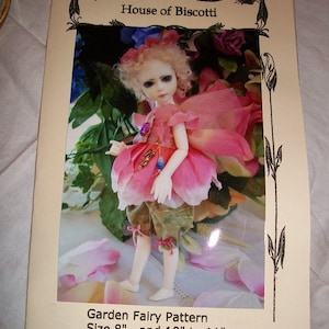 May include: A pink and green fairy doll with a flower crown and wings. The doll is standing on a white surface with pink flower petals. The image is from a pattern for a garden fairy doll, sizes 8 inches and 10 to 11 inches. The text "House of Biscotti" is at the top of the image.
