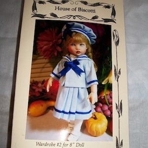 May include: A doll wearing a blue and white sailor dress with a blue ribbon and a blue and white sailor hat. The doll is standing in front of a pumpkin and grapes. The image is from a pattern book for making doll clothes. The text on the page reads "Wardrobe #2 for 8" Doll 3 Dresses, Jacket, 4 Hats, and a Romper".