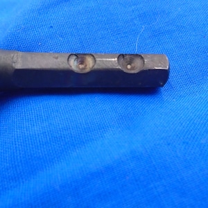 Puede incluir: Primer plano de una herramienta de metal con dos agujeros perforados. La herramienta es de color gris oscuro y tiene una superficie lisa.