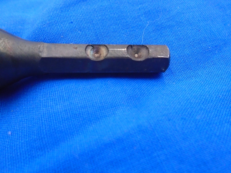 Puede incluir: Primer plano de una herramienta de metal con dos agujeros perforados. La herramienta es de color gris oscuro y tiene una superficie lisa.
