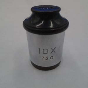 May include: A cylindrical, silver-colored microscope eyepiece with a black top and base. The text "10X 730" is printed on the side. The top has a dark blue interior.