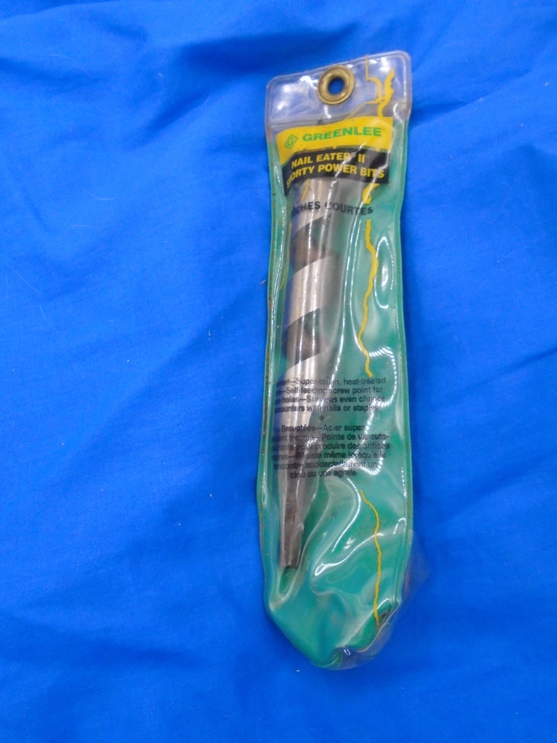 Puede incluir: Un paquete verde y amarillo que contiene una broca Greenlee Nail Eater II de 1/4 de pulgada. El paquete tiene el texto "Greenlee Nail Eater II 1/4 inch Power Bits" e "Inches Courtes" impreso en &eacute;l.
