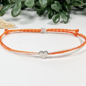 May include: A simple orange and white string bracelet with a silver heart charm.