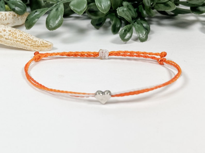May include: A simple orange and white string bracelet with a silver heart charm.