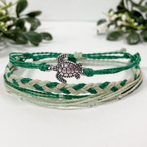 May include: A multi-layered bracelet with a silver turtle charm. The bracelet is made of green, beige, and white string.