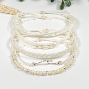 May include: A set of six white string bracelets with silver beads and a silver wave charm. The bracelets are stacked on top of each other.