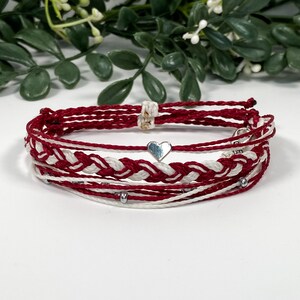 May include: A red and white braided friendship bracelet with a silver heart charm. The bracelet is made of multiple strands of cord and has silver beads.