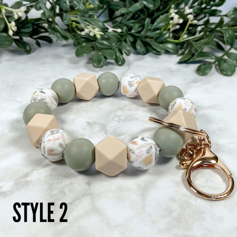 May include: A green and beige silicone beaded keyring with a floral pattern and a gold metal key ring. The keyring is labelled "STYLE 2".