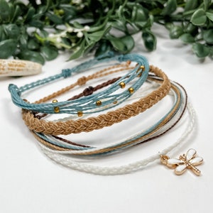 May include: A set of six colorful friendship bracelets with a gold dragonfly charm. The bracelets are made of braided and knotted string in shades of blue, brown, and white.