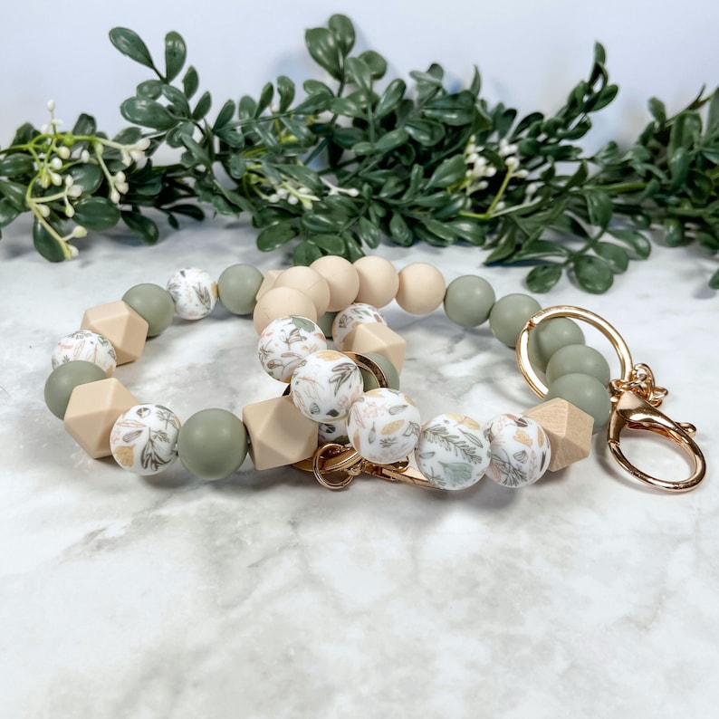 May include: A green and beige silicone beaded keyring with a floral pattern. The keyring has a gold metal clasp.