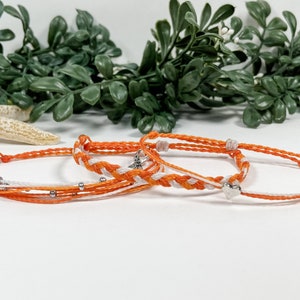 May include: Three friendship bracelets in orange and white with silver beads and a silver charm. The bracelets are made of string and are tied together in a knot.