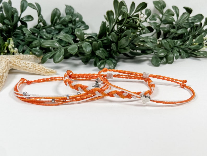 May include: Three friendship bracelets in orange and white with silver beads and a silver charm. The bracelets are made of string and are tied together in a knot.