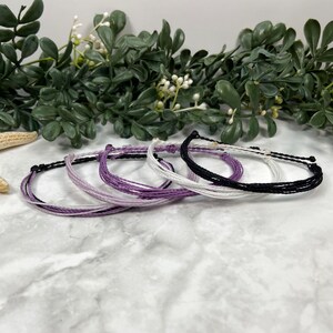 May include: Five colorful friendship bracelets in shades of purple, white, and black. The bracelets are made of thin, braided cords and are tied together in a knot.