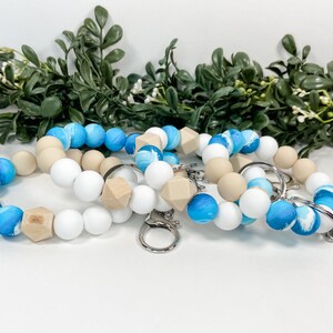 May include: Three silicone beaded keychains with a blue and white marbled design. Each keychain has a wooden bead and a silver key ring.