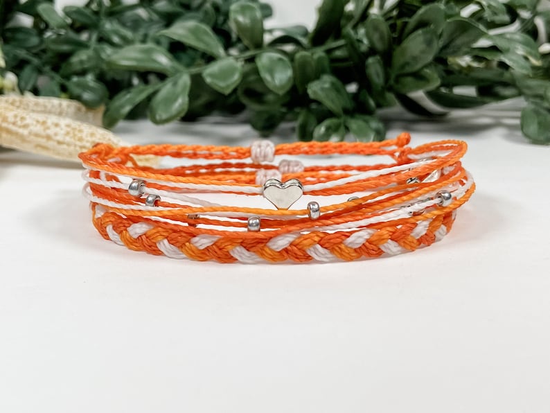 May include: A set of orange and white friendship bracelets with silver heart and bead accents.