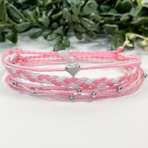 May include: A set of pink and white friendship bracelets with a silver heart charm. The bracelets are made of thin string and are tied together with a knot.