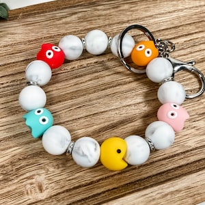 May include: A white marble bead keychain with a silver key ring and colorful Pac-Man character charms. The charms are red, blue, pink, yellow, and orange.