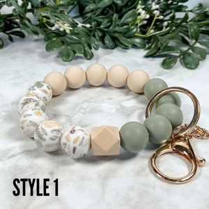 May include: A beige and green silicone beaded keyring with a gold metal key ring. The beads have a floral pattern and a wood bead.